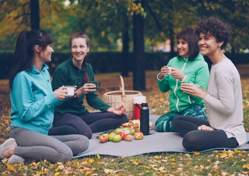 Happy girls are having picnic in park sitting on yoga mats and eating after outdoor practice in autumn, girls are talking and laughing. Communication, people and food concept.