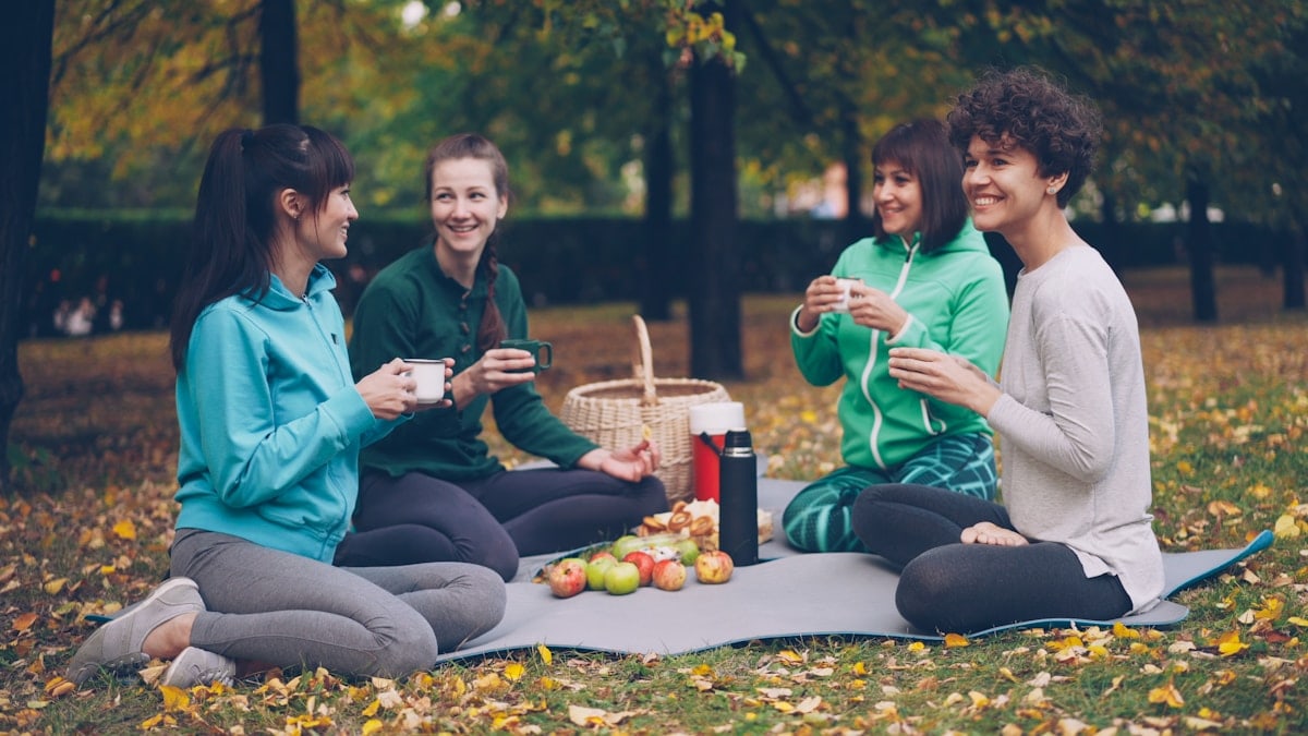 Happy girls are having picnic in park sitting on yoga mats and eating after outdoor practice in autumn, girls are talking and laughing. Communication, people and food concept.