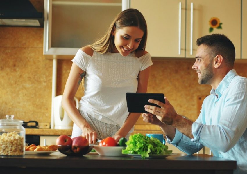Attractive couple chatting in the kitchen early morning. Handsome man using tablet while his girlfriend cooking breakfast at home